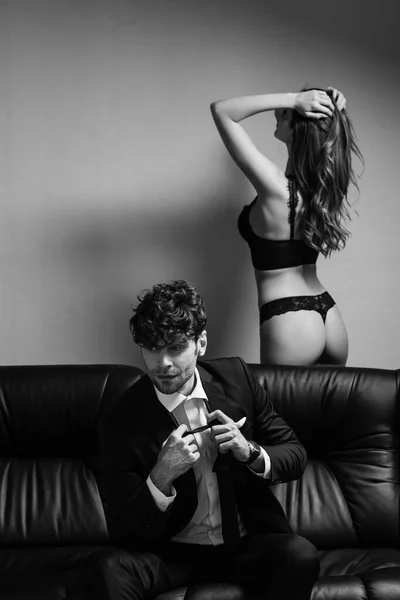 Monochrome image of handsome man in suit holding tie on couch near sexy woman in lingerie on grey background