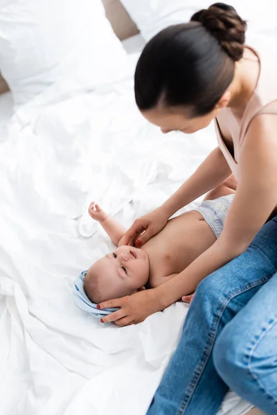 ベッドに座って可愛い赤ちゃんの息子を見ている母親の頭上には — ストック写真