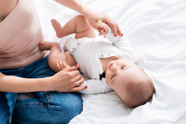 selective focus of mother holding hands with infant son in baby romper on bed