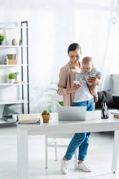 Freelancer Holding Arms Infant Son Using Smartphone Laptop — Stock Photo, Image