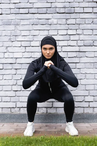 若いイスラム教徒のスポーツ女性がレンガの壁の近くでスクワットをして — ストック写真