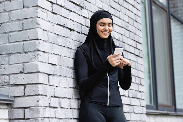happy muslim woman using smartphone and standing near brick wall 