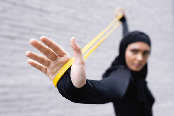 selective focus of arabian sportswoman in hijab exercising with resistance band near brick wall