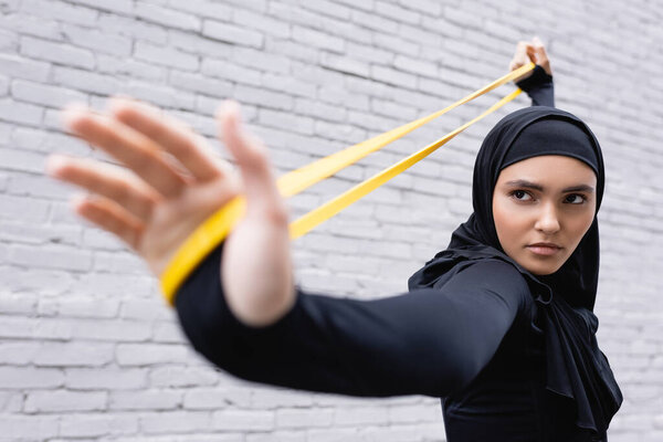 selective focus of arabian woman in hijab exercising with resistance band near brick wall