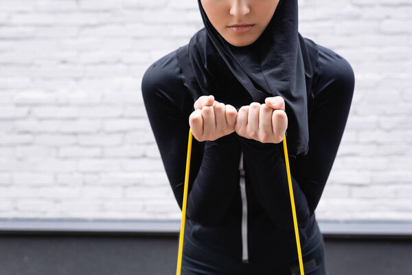 cropped view of arabian sportswoman in hijab exercising with resistance band near brick wall