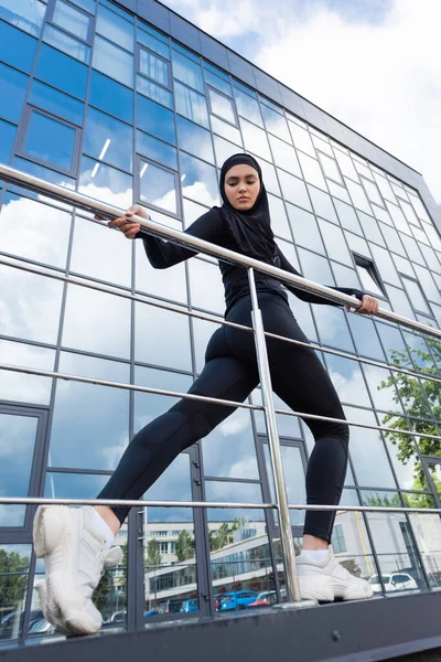 low angle view of arabian woman in hijab holding handrail while working out near modern building