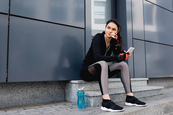 Joven Deportista Sentada Cerca Botella Deportiva Sosteniendo Teléfono Inteligente — Foto de Stock