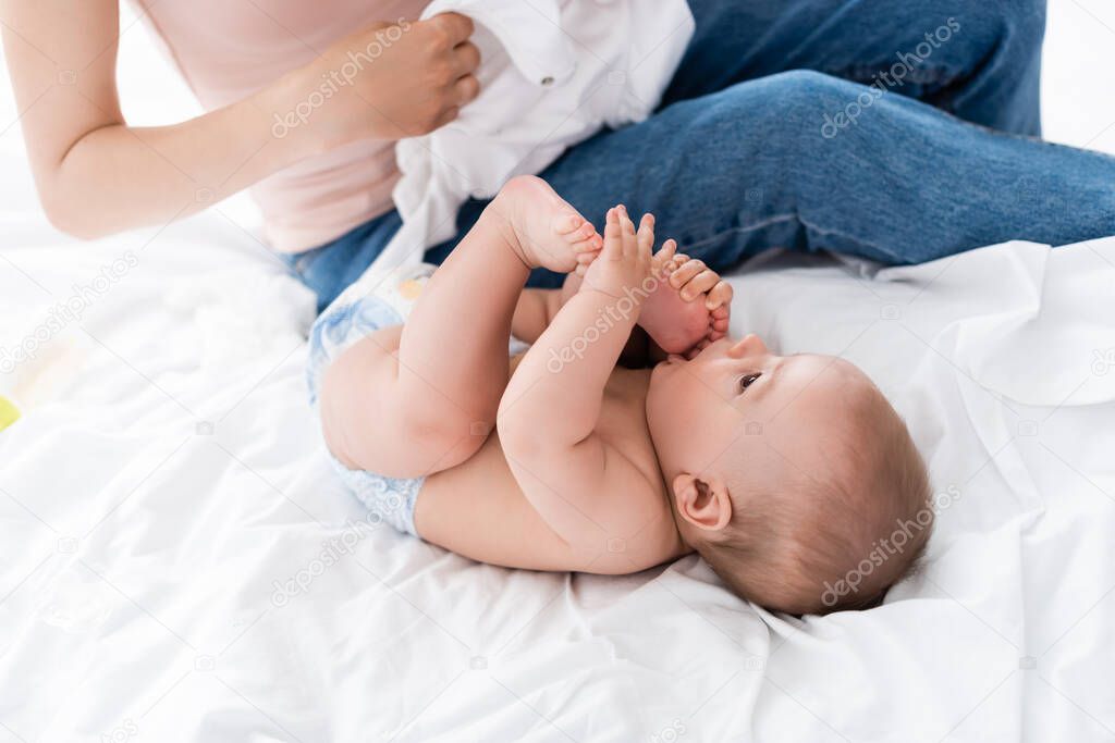 mother sitting on bed and holding baby romper near cute infant son in diaper 