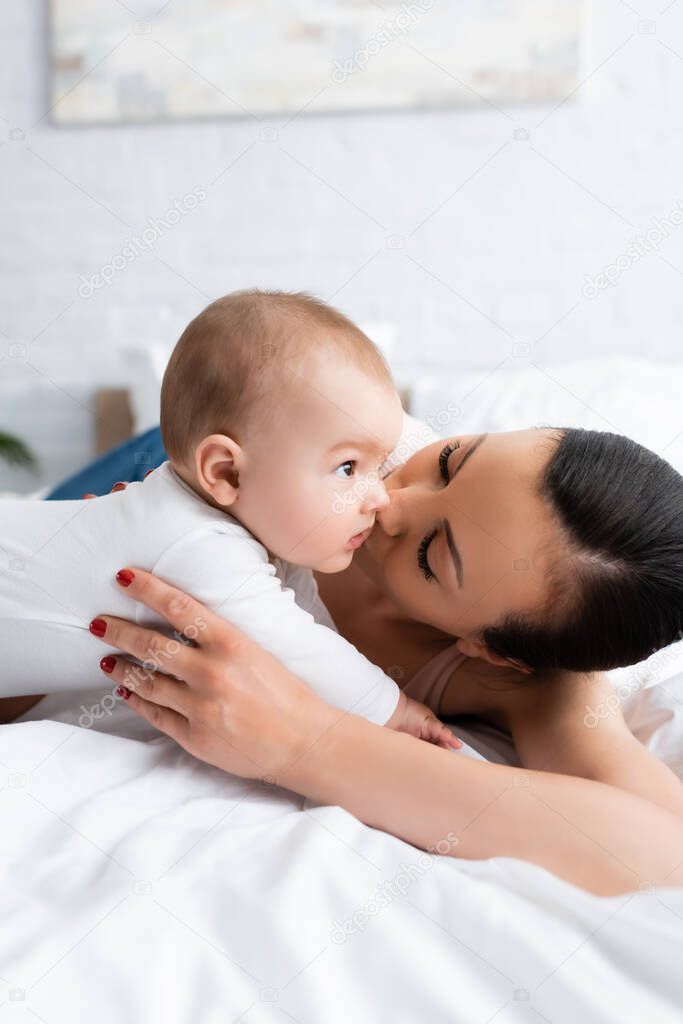 woman kissing cute infant son in baby romper lying on bed