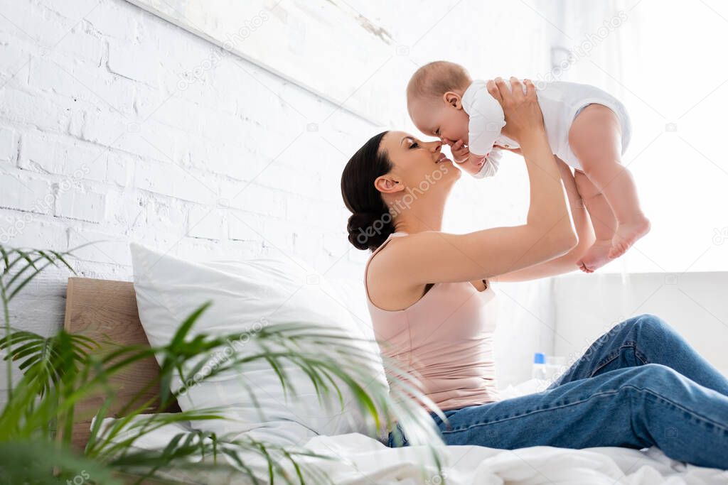 selective focus of cheerful mother in jeans sitting on bed and holding in arms cute infant son 