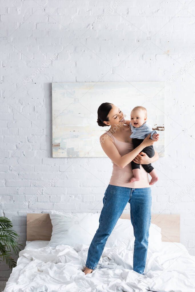 happy mother holding in arms happy barefoot infant son in baby clothing and standing on bed