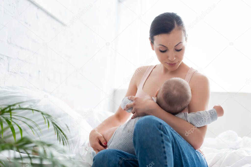 selective focus of attractive mother looking at infant son while breastfeeding in bedroom