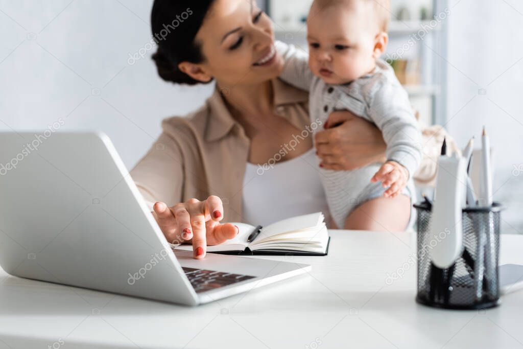 selective focus of happy freelancer holding in arms infant son and using laptop 