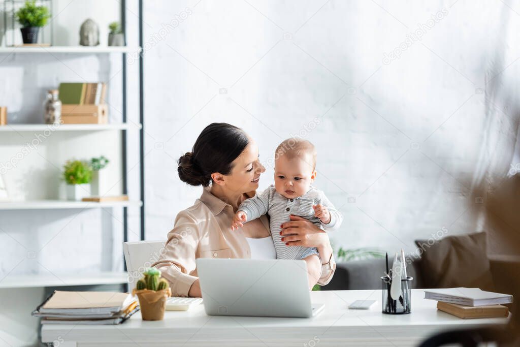 selective focus of happy mother looking at infant son near laptop on table