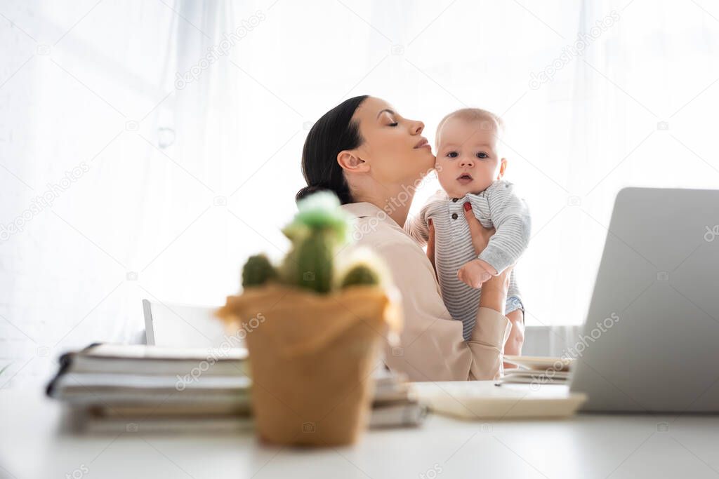 selective focus of mother with closed eyes holding in arms cute infant son near laptop and cactus on table