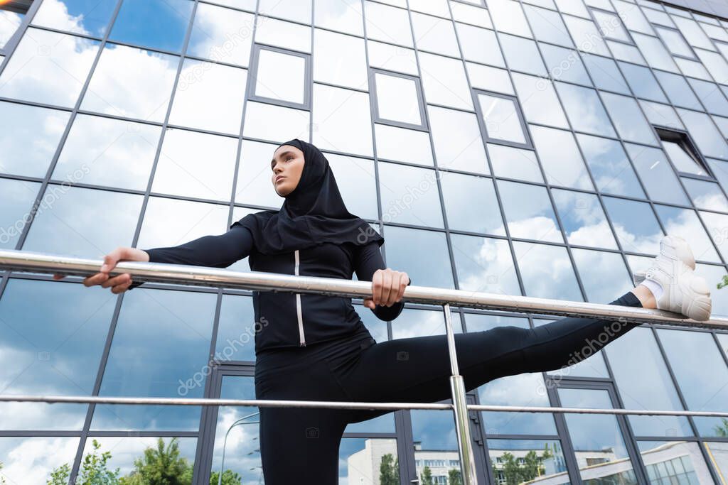 low angle view of arabian woman in hijab holding handrail while exercising near modern building 