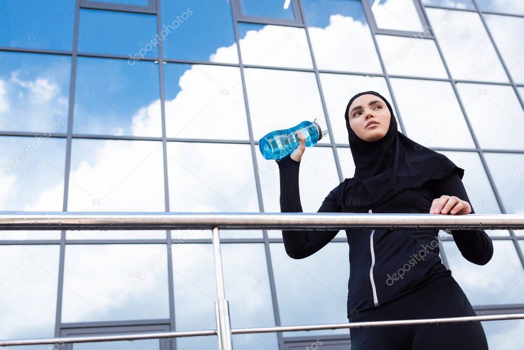 low angle view of young muslim woman in hijab holding sports bottle near modern building 