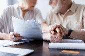 Oříznutý pohled na starší pár držící se za ruce u dokumentů, notebooku a kreditní karty na stole 