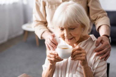 Selective focus of senior man embracing wife with cup of coffee at home  clipart