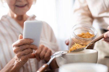 Kocası mısır gevreğini kaseye dökerken yaşlı bir kadının akıllı telefon kullandığı seçici bir odak noktası.