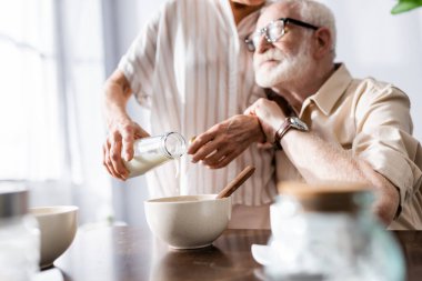 Selective focus of man hugging senor wife pouring milk in bowl at table  clipart