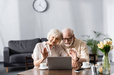 Evde dizüstü bilgisayar başında gülümseyen yaşlı çiftin el sallamasının seçici odağı. 