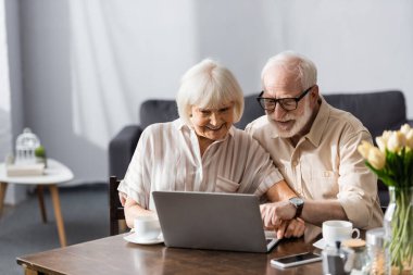 Masadaki kahve fincanlarının yanında dizüstü bilgisayar kullanarak gülümseyen yaşlı çiftin seçici odağı 