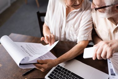 Elinde kâğıtları olan, akıllı telefonlu, gülümseyen yaşlı bir kadının yüksek açılı görüntüsü 
