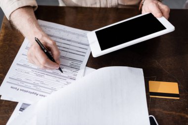 Son sınıf öğrencisinin kağıtlara yazı yazması ve kredi kartının yanında dijital tablet tutması. 