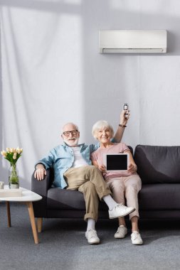 Gülümseyen yaşlı çift dijital tablet gösteriyor ve evde uzaktan kumandalı klima tutuyor. 