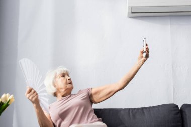 Yaşlı bir kadının yelpaze tutarken ve uzaktan kumandalı klima kullanırken seçici odak noktası 