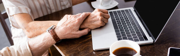 Panoramic crop of senior man holding hand of wife near gadgets and coffee cups on table 