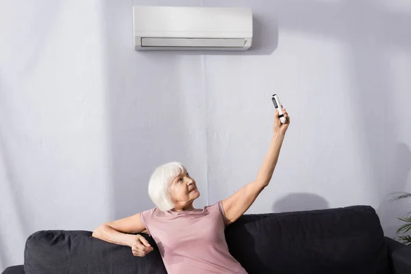 リビングルームでソファの上にエアコンのリモコンを持っている高齢女性 — ストック写真