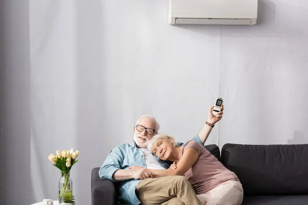 Ηλικιωμένη Γυναίκα Αγκαλιάζει Χαμογελαστός Σύζυγος Τηλεχειριστήριο Του Κλιματιστικού Στο Σπίτι — Φωτογραφία Αρχείου