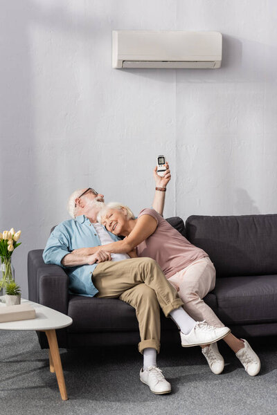 Smiling elderly woman embracing husband using remote controller of air conditioner in living room
