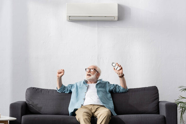 Senior man showing yes gesture while holding remote controller under air conditioner on sofa 