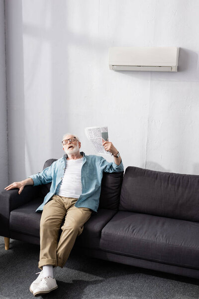 Старший мужчина размахивает газетой, страдая от жары на диване
 