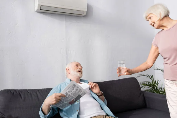 Starší Žena Dává Sklenici Vody Manželovi Drží Noviny Pod Klimatizací — Stock fotografie