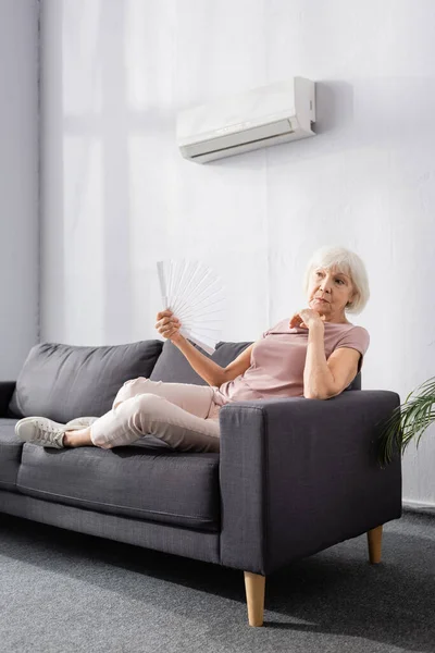 Elderly woman waving fan under air conditioner in living room — Stock Image Elderly Woman Waving Fan Air Conditioner Living Room Stock Image