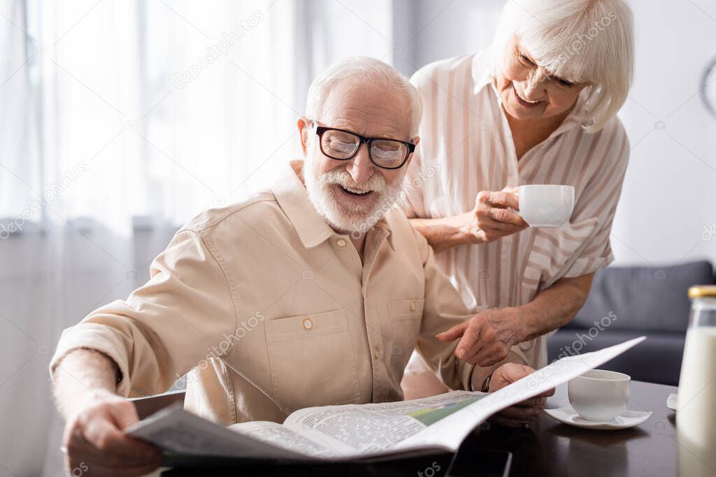 Selective focus of smiling senior man reading newspaper near wife with cup of coffee in kitchen  