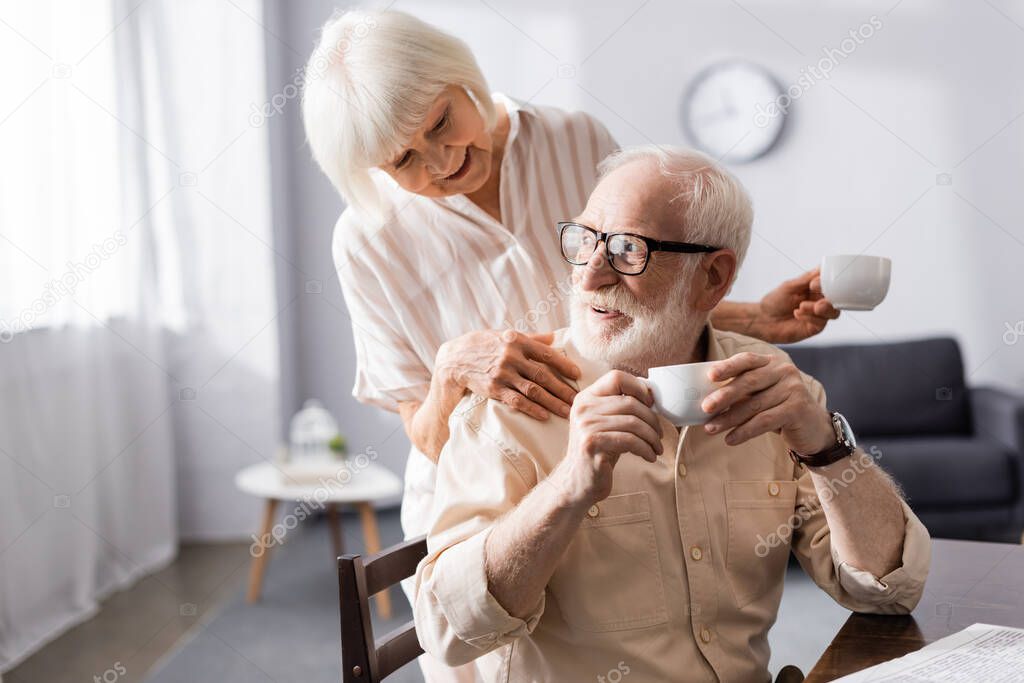Smiling senior woman holding cup of coffee and embracing husband during breakfast at home 