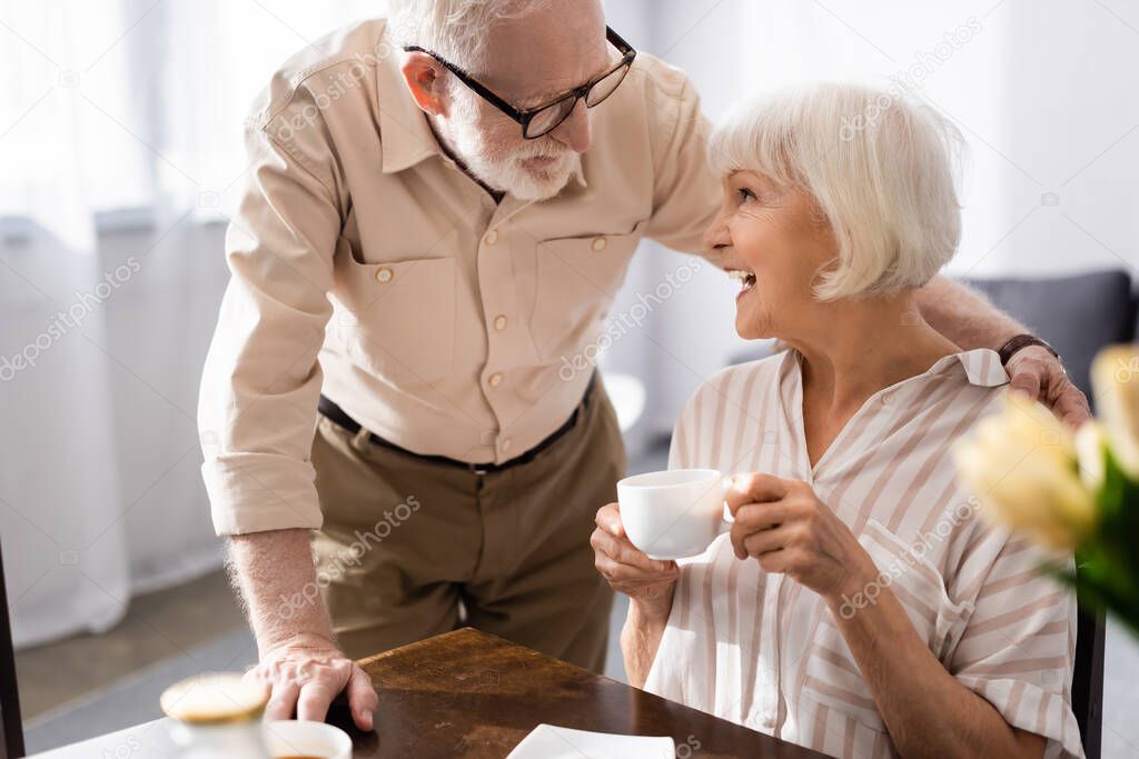 Selective focus of senior man hugging positive wife with cup of coffee in kitchen 