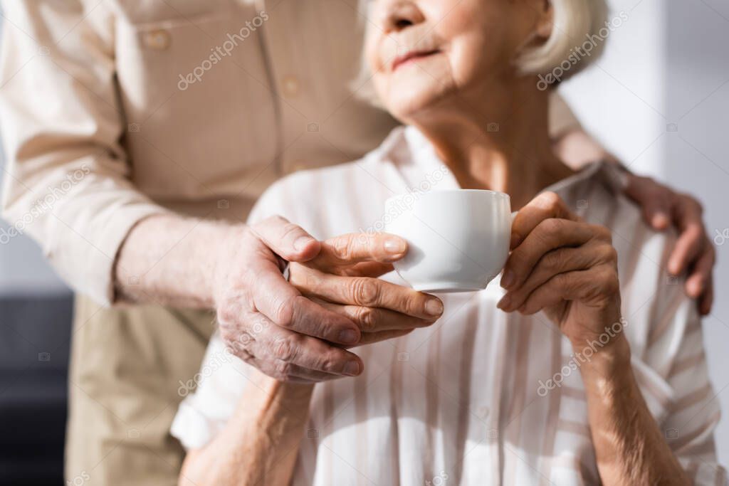 Cropped view of senior man embracing wife holding coffee cup at home 