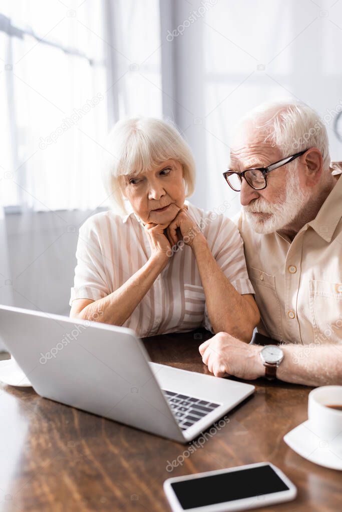 Selective focus of elderly couple looking at laptop near smartphone and coffee on table 