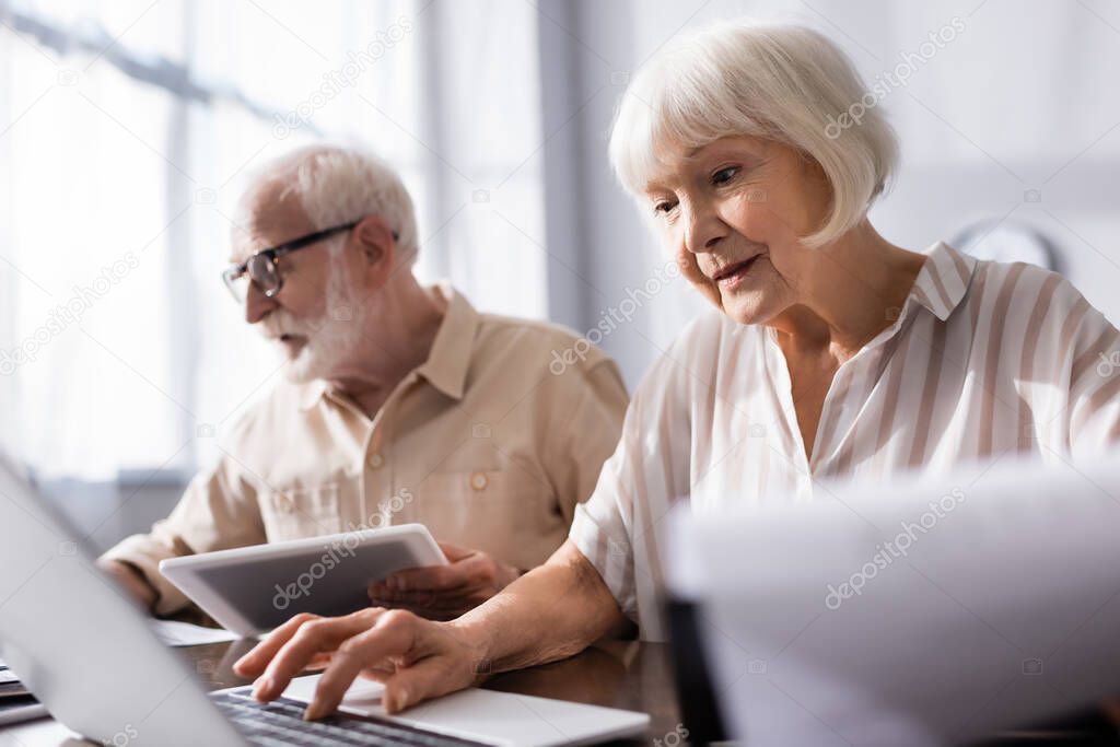 Selective focus of senior couple using gadgets near papers on table at home 
