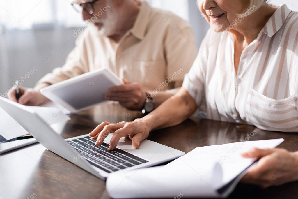 Selective focus of elderly woman using laptop and holding papers near husband with digital tablet at home 