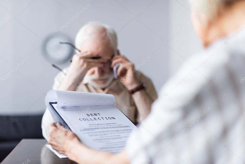Selective focus of woman holding documents with debt collection lettering while sad husband talking on smartphone 
