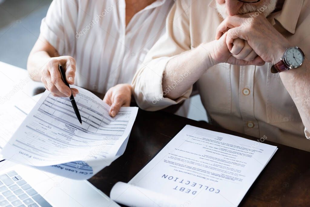 Cropped view of senior woman pointing at papers near husband and document with debt collection lettering on table  