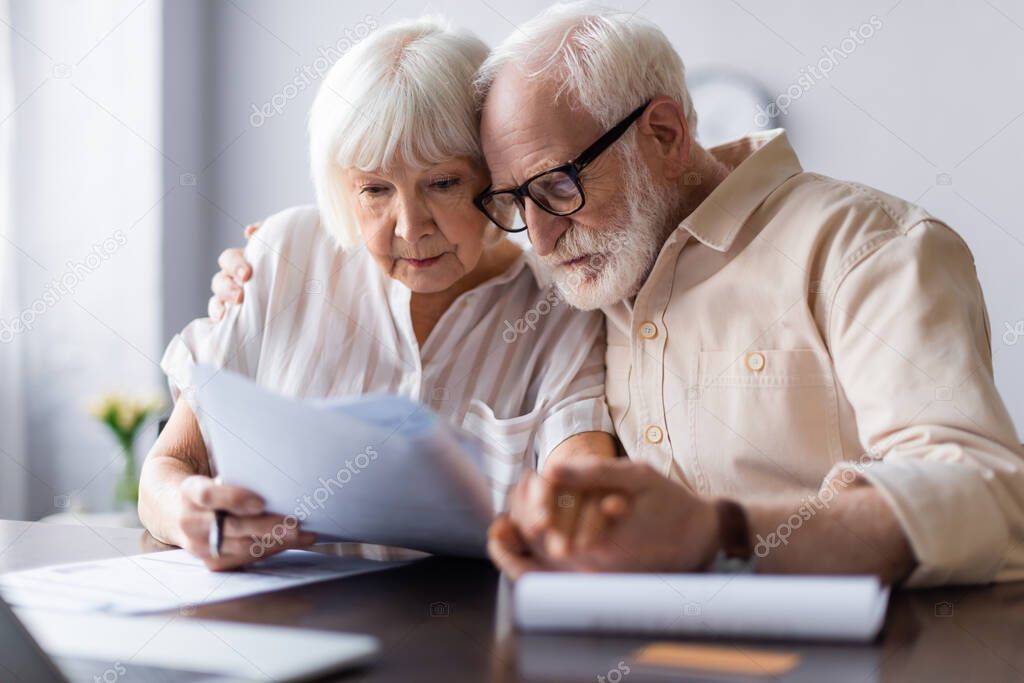 Selective focus of senior man embracing wife holding papers at home 