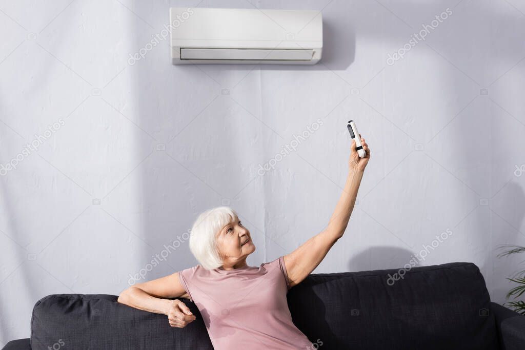 Elderly woman holding remote controller of air conditioner on couch in living room